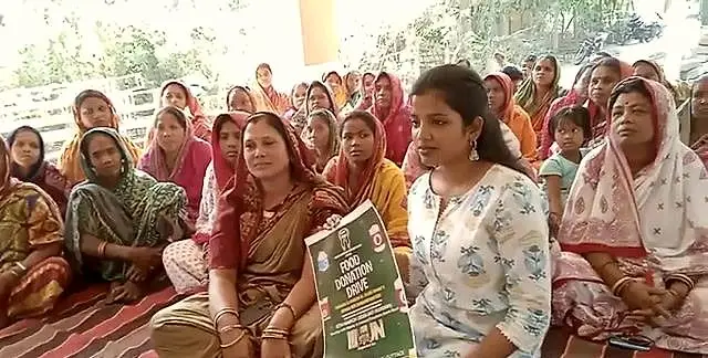 Women sit together on mats at a WCWS community programme, one holding an awareness poster.